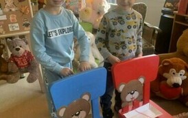 Dw&oacute;ch chłopc&oacute;w stoi przy dw&oacute;ch krzesłach na kt&oacute;rych oparciach są naklejone głowy misi&oacute;w, a na siedziskach lezą talerzyki papierowe z rozwiniętą wł&oacute;czką. Dzieci z tyłu krzeseł nawijają wł&oacute;czkę na rolkę, ktora przechodzi przez usta misia wizualizując jedzen