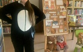 Pani stoi w białoczarnym przebraniu misia i się uśmiecha. Po prawej stronie stoi korkowa tablica z grafiką i informacjami o Dniu Misia.