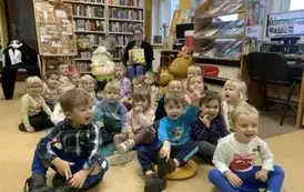 Dzieci siedzą na siedziskach, a z tyłu siedzi Pani bibliotekarka z książką w rękach.