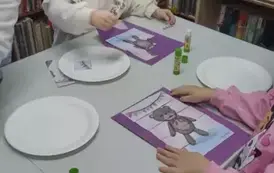 Dwie dziewczynki przy stole naklejają misiowe puzzle na kolorowe kartki. Na stole leżą papierowe talerzyki na kt&oacute;rych były papierowe puzzle.