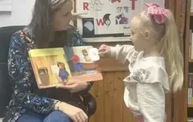 Bibliotekarka ma otwartą książkę i stoi przy Niej dziewczynka w blond włosach i otwiera okienko w książeczce.