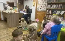 Dzieci siedzą na siedziskach i na krzesełku i słuchają bajki, kt&oacute;rą czyta im bibliotekarka pokazując im obrazki w książce.