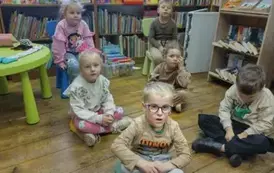 Dzieci siedzą na siedziskach i słuchają bajki, kt&oacute;rą czyta im bibliotekarka.