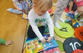 Dziewczynka w blond włosach bawi się pluszową książeczką, obok są inne dzieci.