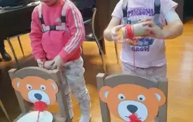 Dwie dziewczynki stoją za dwoma krzesełkami na kt&oacute;rych oparciach naklejone są twarze misi&oacute;w, a na siedzeniach są położone talerzyki papierowe z rozwiniętą wł&oacute;czką, kt&oacute;ra przechodzi przez usta misia do rąk dzeci , kt&oacute;re tą wł&oacute;czkę nawijają na rolkę .