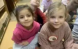 Z przodu siedzą dwie dziewczynki, jedna w jasnych, a druga w ciemnych włosach. Za nimi siedzą inne dzieci.