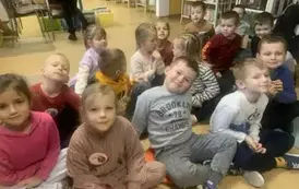 Dzieci siedzą na siedziskach w bibliotece i słuchają bajki.