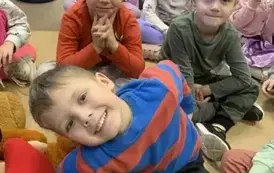 Dzieci siedzą na siedziskach, z przodu chłopiec siedzi pochylony w lewą stronę i się uśmiecha.