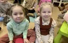 Dwie dziewczynki siedzą z przodu na siedziskach i się uśmiechają. Za nimi widać inne dzieci.