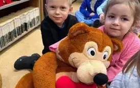 Dziewczynka w blond włosach siedzi koło brązowego misia i go przytula do siebie. Za nią widać inne dzieci.