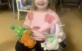 Uśmiechnięta dziewczynka siedzi na podłodze i trzyma książeczki pluszowe w kształcie maskotek.