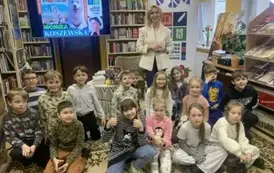 Dzieci w grupie siedzą na dywanie, a za nimi stoi pisarka w białej marynarce. Jedna z dziewczynek ma podniesione dwa kciuki w g&oacute;rę.