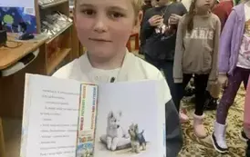 Chłopiec w blond włosach pokazuje swoją nową otwartą książkę z zakładką w środku. W tle są inne dzieci.