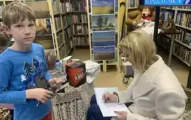 Chłopiec w niebieskiej bluzce stoi przy pisarce, kt&oacute;ra mu podpisuje zakupioną książkę.