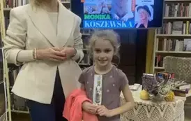 Dziewczynka w blond włosach stoi przy pisarce, ma na ręce przewieszoną bluzę i trzyma w rękach zakładkę do książki. Obie się uśmiechają.