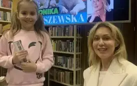 Uśmiechnięta dziewczynka w dw&oacute;ch kitkach stoi przy pisarce, kt&oacute;ra podpisuje jej zakupioną książkę. 