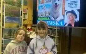 Dwie dziewczynki w siwych bluzach stoją przy pisarce, kt&oacute;ra podpisuje zakupione książki. Nad nimi wisi duży telewizor z wyświetlanym plakatem dot. tego spotkania.