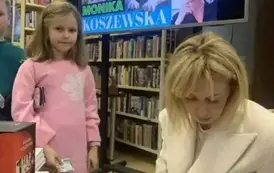 Dziewczynka w r&oacute;żowej bluzce stoi przy pisarce, kt&oacute;ra podpisuje jej zakupioną książkę. U g&oacute;ry wisi telewizor z wyświetlanym plakatem dot. tego spotkania.