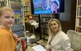 Chłopiec w pomarańczowej bluzie stoi przy pisarce, kt&oacute;ra podpisuje mu książkę, kt&oacute;rą kupił.