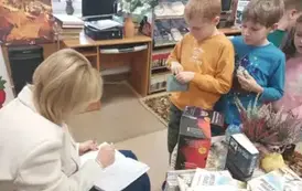 Pisarka przy stoliku sprzedaje swoje książki i dzieci podchodzą kolejno po jej zakup i autograf.