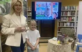 Pisarka stoi przy dziewczynce w blond włosach, kt&oacute;ra się ładnie uśmiecha.