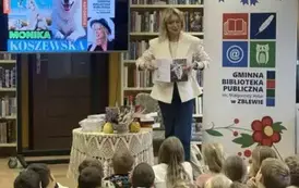 Pisarka stoi przy stoliku i pokazuje okładkę swojej książki. Po lewej stronie wisi telewizor z wyświetlanym plakatem dot. spotkania,a po prawej stronie stoi rolap z logiem biblioteki.