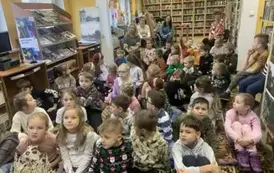 Dzieci siedzą w dużej grupie na dywanie, a z tyłu na krzesłach siedzą nauczyciele.