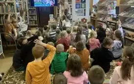 Dzieci siedzą na dywanie i niekt&oacute;re z nich podnoszą ręce, a z przodu pisarka pochylona jest nad stolikiem. 