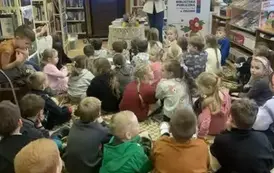 Dzieci siedzą na dywanie,a z przodu stoi Pisarka i opowiada. Trzyma w ręku swoje książki. Po lewej stronie wisi telewizor z wyświetlanym plakatem dot. tego spotkania, a po prawej stronie stoi rolap z logiem biblioteki.