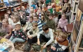 Duża grupa dzieci siedzi na dywanie, a za nimi siedzą na krzesłach nauczyciele.