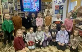 Zdjęcie grupowe dzieci z pisarką, kt&oacute;ra stoi za nimi. Po lewej strronie jest telewizor z wyświetlanym plakatem dot. spotkania,a po prawej stronie jest rolap z logiem biblioteki.