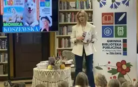 Pisarka w białej marynarce stoi przy stoliku i trzyma kilka swoich książek. Po lewej stronie wisi telewizor z wyświetlanym plakatem dotyczącym tego spotkania. Na stoliku leżą książki autorki, kwiat i małe dynie.