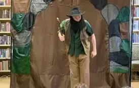 Na tle wejścia do jaskini stoi pochylony aktor przebrany w brązowozielone ubranie w czarnej brodzie i kapeluszu z laską w ręku.