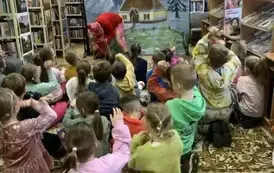 Dzieci siedzą w grupie na dywanie, niekt&oacute;re z nich podnoszą rękę. Z przodu na tle chaty zimą w g&oacute;rach aktor przebrany za renifera pochylony jest nas sankami i trzyma je w ręku.