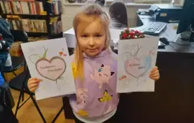 Dziewczynka w dłuższych blond włosach pokazuje swoje dwie laurki - dla babci i dziadka.