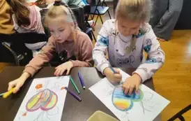 Dwie dziewczynki w blond włosach przy stole kolorują ładnie laurki dla babci i dziadka. Z tyłu widać przy stole inne dzieci.