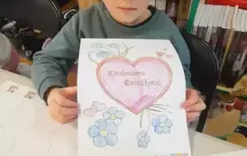 Chłopiec w blond włosach pokazuje przy stole swoją laurkę dla dziadka. Obok na stole leżą inne laurki.