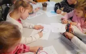 Dzieci siedzą przy białym stole i kolorują kredkami laurki dla babci i dziadka