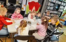 Dzieci siedzą przy białym stole i kolorują kredkami laurki dla babci i dziadka