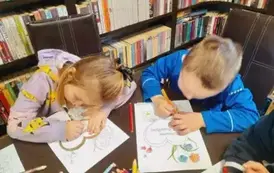 Dziewczynka z chłopcem przy stole kolorują kredkami laurki dla babci i dziadka