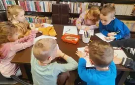 Dzieci siedzą przy stole i kolorują kredkami laurki dla babci i dziadka