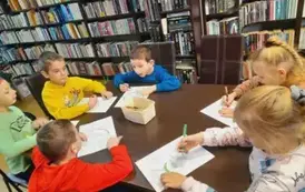 Dzieci siedzą przy stole i kolorują kredkami laurki dla babci i dziadka