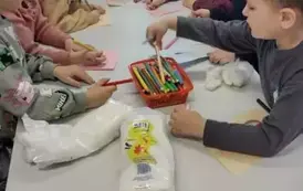 Dzieci przy białym stole przystrajają ma kartkach bałwanki za pomocą kredek, krepy i waty. Nad nimi pochyla się nauczycielka i im pomaga.