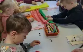 Dzieci przy białym stole przystrajają ma kartkach bałwanki za pomocą kredek, krepy i waty.