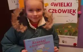 Dziewczynka w blond włosach trzyma swoją nową książeczkę z wyprawki .Za nią stoi tablica z plakatem projektu.
