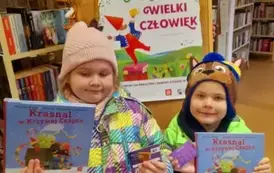 Brat z siostrą stoją obok siebie i trzymają nowe książeczki z wyprawki czytelniczej i karty Małego Czytelnika. Za nimi jest tablica z plakatem dotyczącym projektu