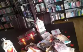 Na stole są wyłożone książki o tematyce miłosnej przykryte lampkami i otoczone serduszkami. Za nimi stoją zapalone dwa lampiony i dwie świeczki.