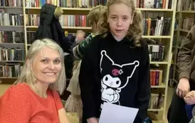 Dziewczynka w blond włosach w czarnej bluzie trzyma białą kartkę, stoi przy stoliku pisarki.