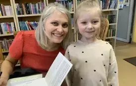 Dziewczynka w blond włosach stoi przy pisarce aby zakupić książeczkę. Obie się uśmiechają. 