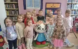 Na tle bajkowego pokoju przy Kici Koci stoi grupa dzieci, kt&oacute;re trzymają Kicię za głowę.